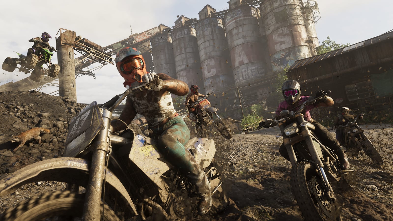 Off-road motorcyclists riding through rough terrain below industrial silos